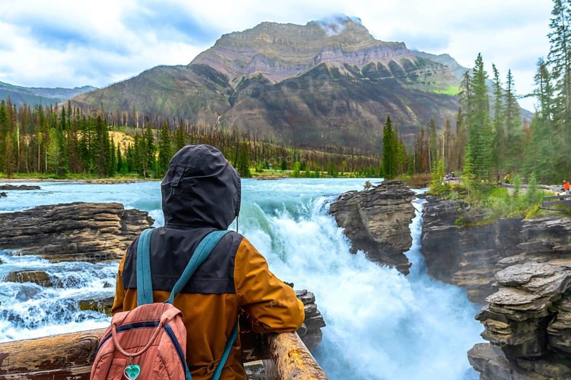 Tourist wearing a backpack and hooded jacket admiring the breathtaking view of powerful athabasca falls in jasper national park, alberta's stunning natural wonder