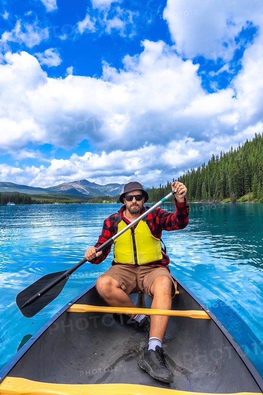 Tourist wearing sunglasses and a life jacket paddling a canoe on turquoise maligne lake under a bright blue sky, surrounded by lush forests and mountains in jasper national park, alberta