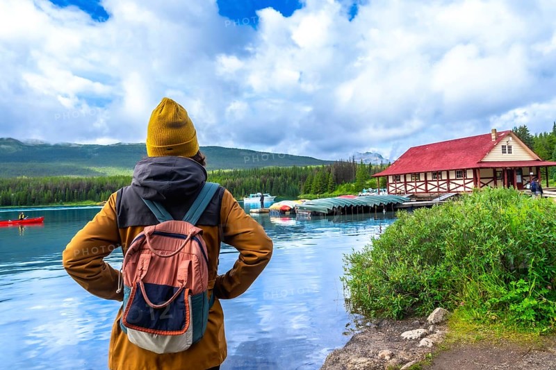Female tourist wearing a backpack standing on the shore of maligne lake, admiring the iconic boathouse and canoes on a beautiful sunny day in jasper national park, alberta, canada