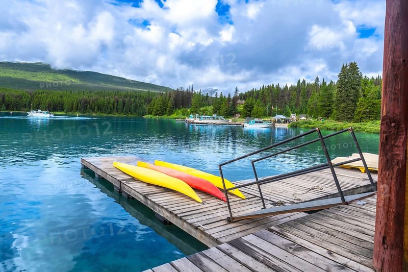 Colorful kayaks lined up on a wooden dock at maligne lake in jasper national park, alberta, while tourist boats navigate turquoise waters beneath a cloudy sky