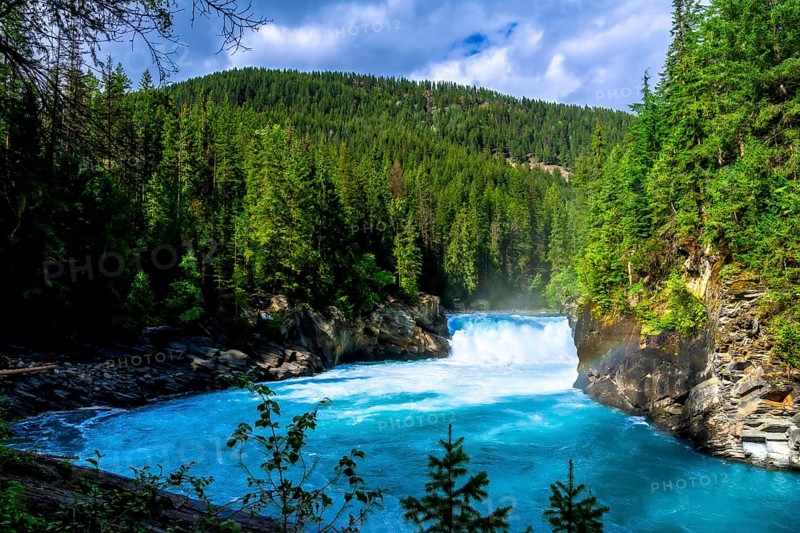Flowing turquoise water of the fraser river winds through a lush green forest toward overlander falls in mount robson provincial park, british columbia, under a cloudy sky