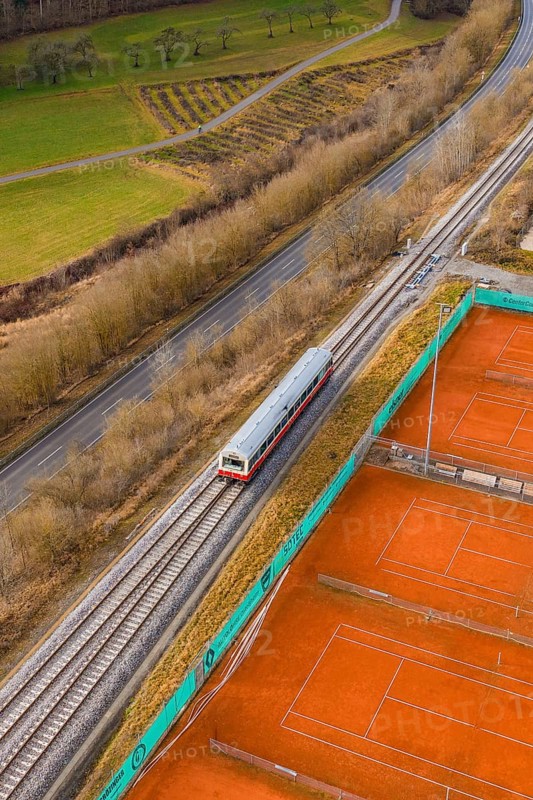A train travels through rural landscape next to orange tennis courts on railroad tracks, historic event, first test run with a passenger train on the Hermann tracks, Hesse Railway since 1988, Calw district, Germany