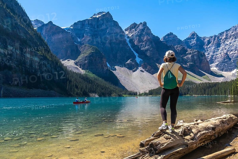 Female tourist standing on a log, admiring turquoise waters of moraine lake with canoers, surrounded by the majestic canadian rockies on a sunny summer day