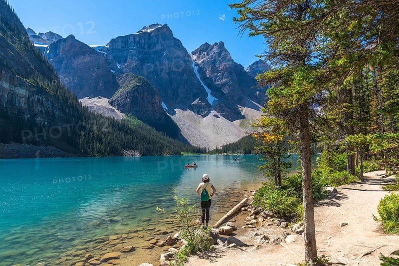 Female tourist standing on the shore of the turquoise moraine lake, admiring the canadian rockies in banff national park on a sunny day with canoes on the water