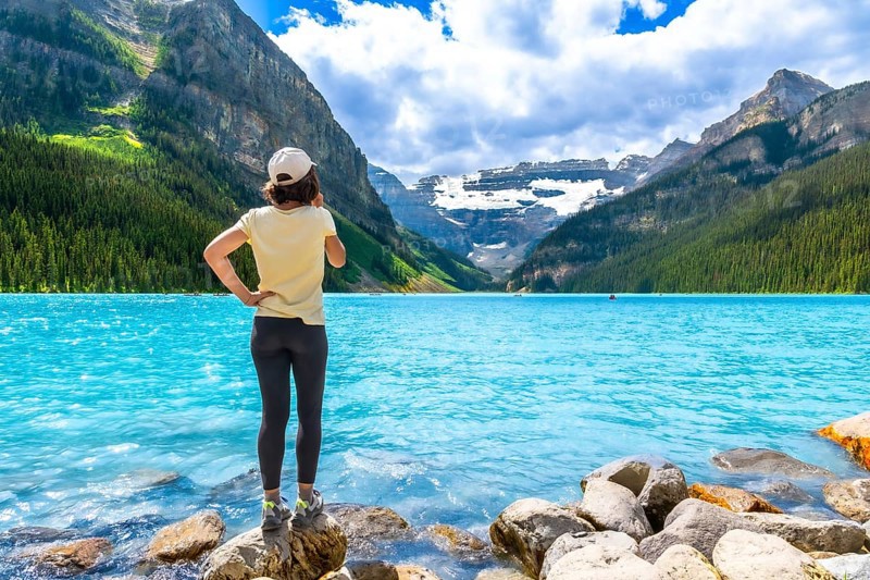 Tourist standing on rocks at the edge of vibrant turquoise waters of lake louise, admiring majestic mountains and glaciers in banff national park on a beautiful summer day