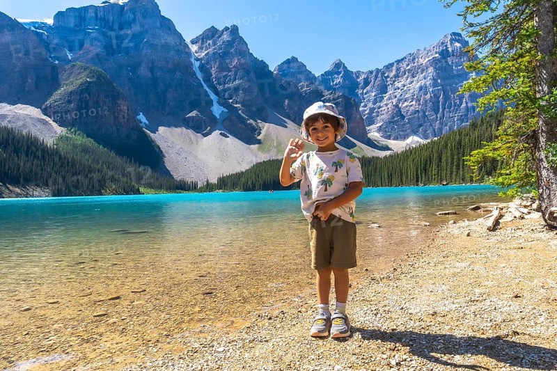 Young boy waving on the shore of moraine lake with the canadian rockies in the background, enjoying a sunny day in banff national park, alberta, canada, during summer vacation