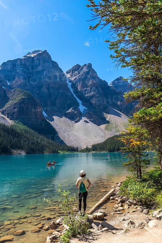 Female tourist standing on the shore of pristine turquoise moraine lake, admiring the majestic canadian rockies in banff national park on a clear summer day