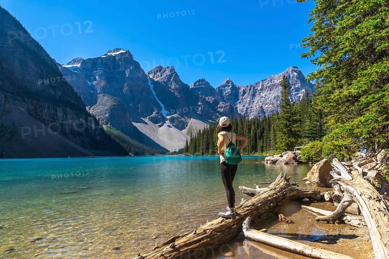 Female tourist standing on a log admiring the turquoise waters of moraine lake with the valley of the ten peaks in the background, banff national park, alberta, canada
