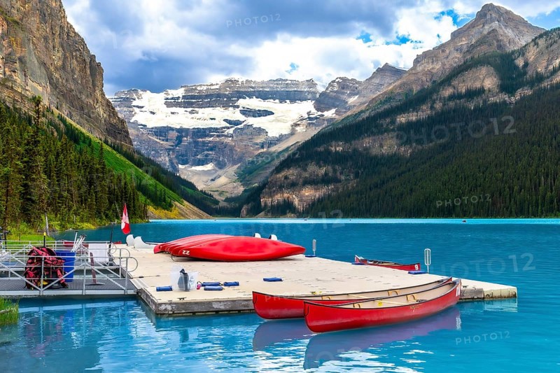 Red canoes are tied to a dock on the turquoise water of lake louise with a backdrop of snow capped mountains and evergreen forest in banff national park, alberta, canada