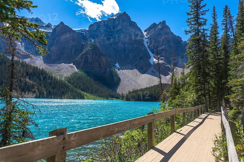 Moraine lake in banff national park features turquoise water, a wooden boardwalk, snow capped mountains, and lush vegetation during a sunny summer day in the canadian rockies