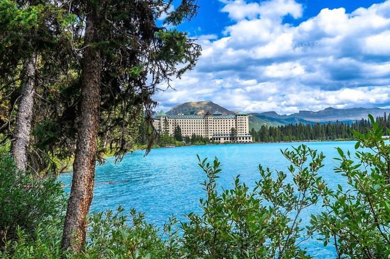 Lush vegetation framing turquoise waters of lake louise reflects a luxury hotel against stunning mountain peaks and a cloudy summer sky in banff national park, alberta