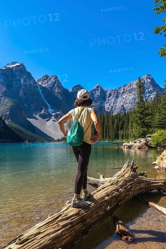 Female tourist standing on a log, admiring the turquoise waters of moraine lake, surrounded by the majestic canadian rockies, in banff national park, alberta, canada, on a sunny summer day