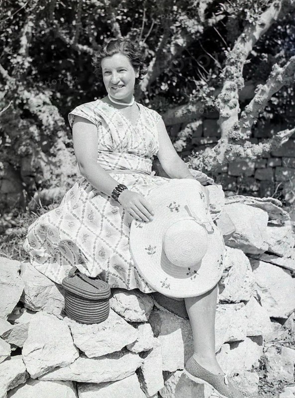 Portrait of young woman wearing summer dress sitting on stone wall holding straw hat, Malta, Europe, 1956