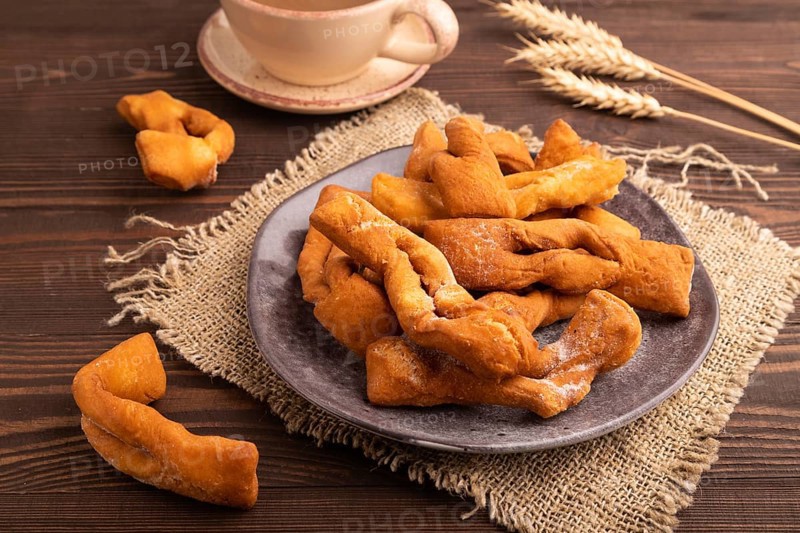 Crunchy biscuit Brushwood cookies sprinkled with powdered sugar on brown wooden background and linen textile, cup of coffee, side view, close up