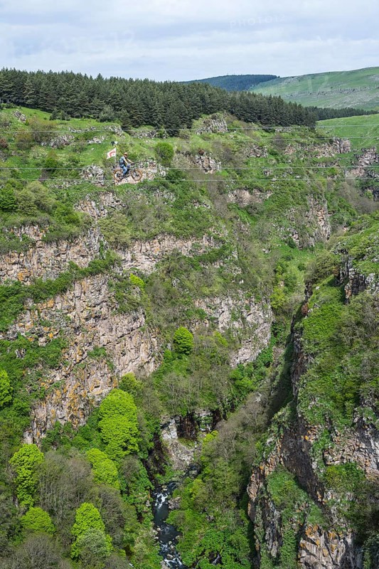 A green gorge with rocks and trees crossed by a small river under a cloudy sky, man riding a bicycle on a stretched rope, Dashbashi Gorge, Khrami River, Chrami, Tsalka Nature Reserve, Kartli, Georgia