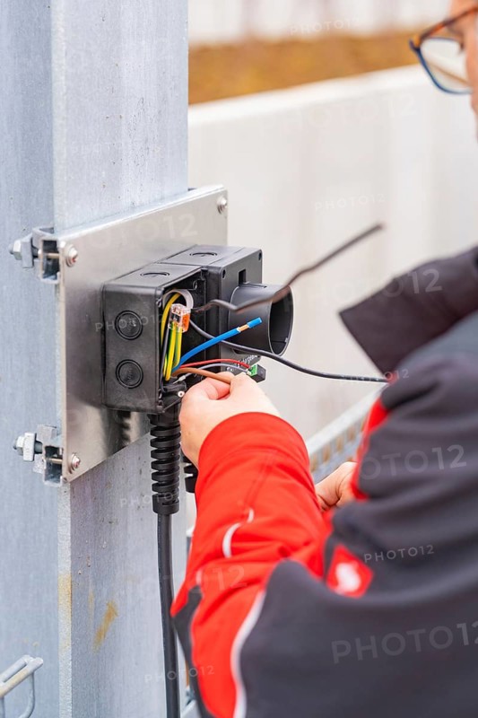Person wearing a red jacket assembles electrical cables at a metal station, parking garage construction with charging infrastructure, Calw, Germany