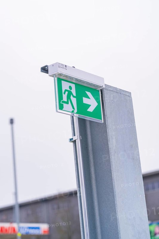 Green emergency exit sign with directional arrow on a metal post, parking garage construction with charging infrastructure, Calw, Germany