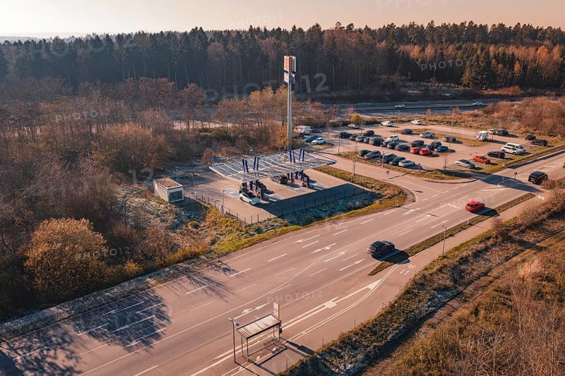 Wide angle view of a street with cars and a charging station, surrounded by autumn trees and parking areas, ENBW Ladepark, Rutesheim, Germany