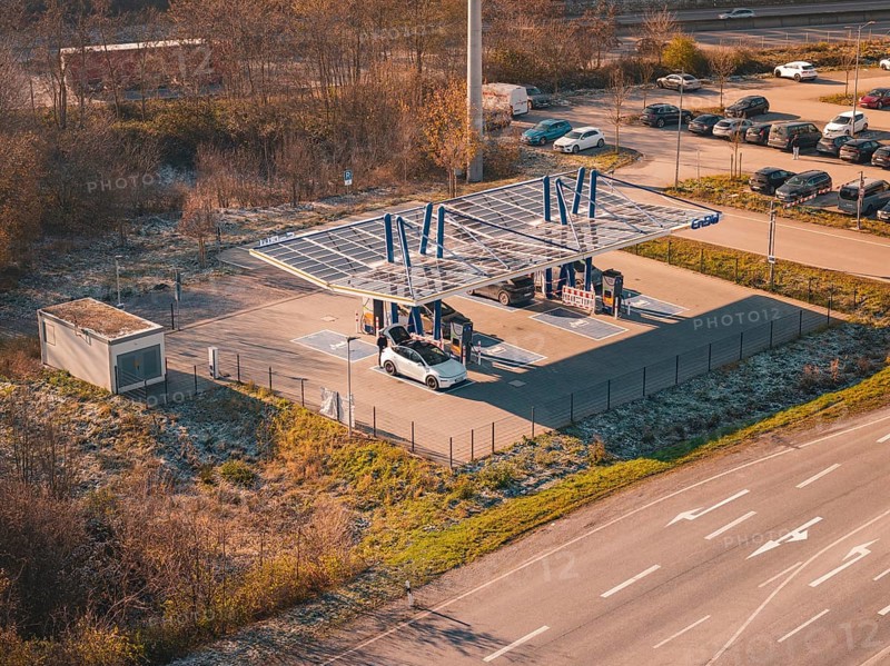 Electric car at a charging station under a solar roof, surrounded by autumn trees and cars in a parking lot, ENBW charging park, Rutesheim, Germany