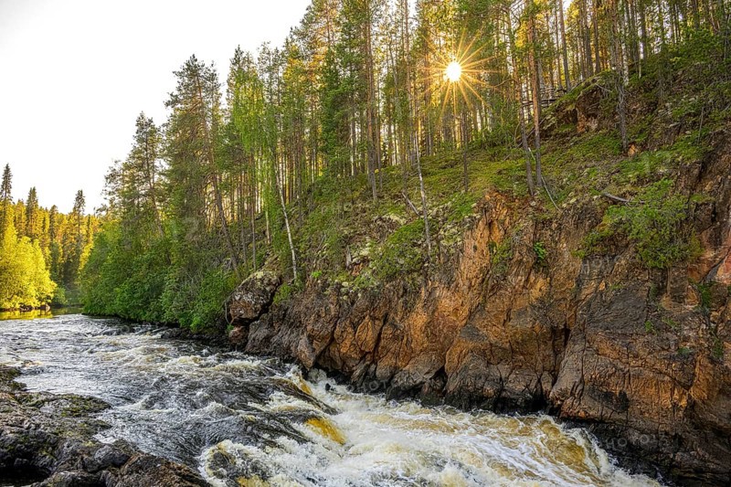A river flows through a thick forest as the sun shines over the treetops and illuminates the rocks, Hossa National Park, Juuma, Kuusamo, Northern Ostrobothnia region, Finland