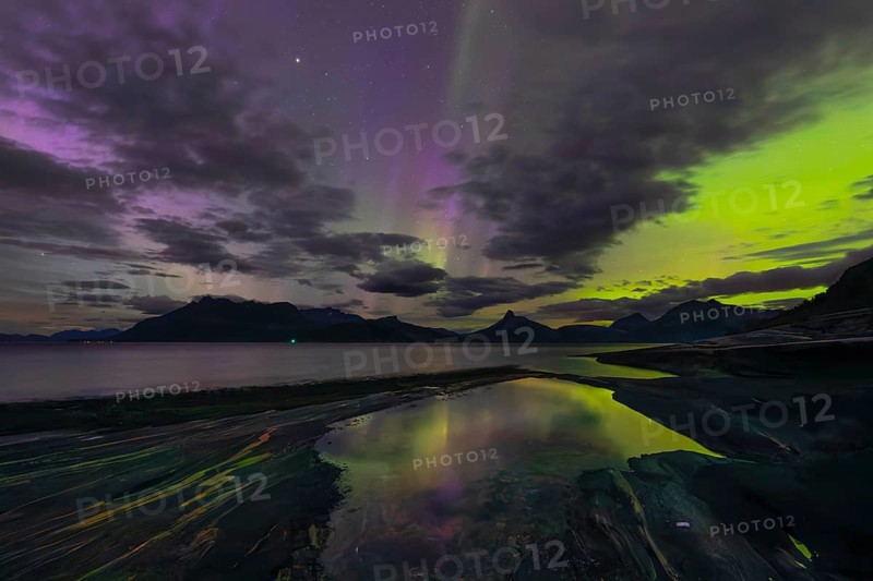 The Vestfjord in Nordland, Norway, has an enchanting atmosphere at night. Dark clouds and vivid aurora, polar lights dance over the fjord. The rugged cliffs and majestic mountains are reflected in the calm water and create a mystical backdrop