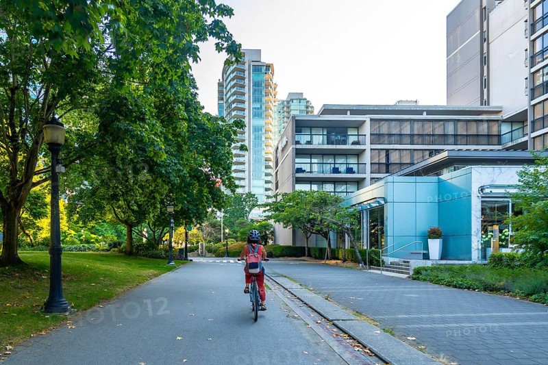 Woman with backpack and helmet is riding a bicycle on a paved path through a green park, passing by modern residential and commercial buildings in vancouver, british columbia