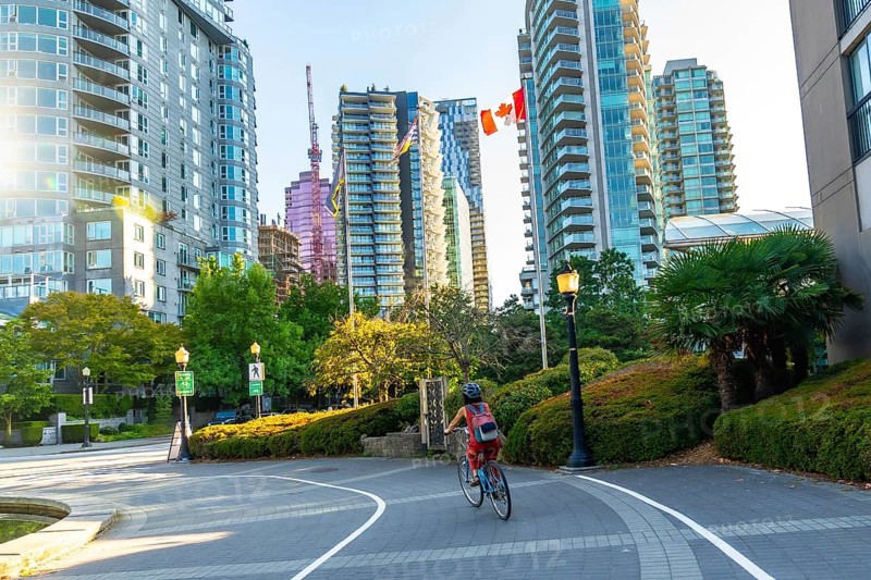 Woman in a helmet and backpack riding a bicycle on a paved path in vancouver, british columbia, surrounded by modern buildings, green trees, and a clear sky