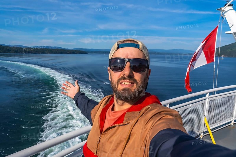 Tourist wearing sunglasses and a cap enjoys ferry trip from victoria, vancouver island, to vancouver, british columbia, opening his arms and showing canadian flag and ocean wake