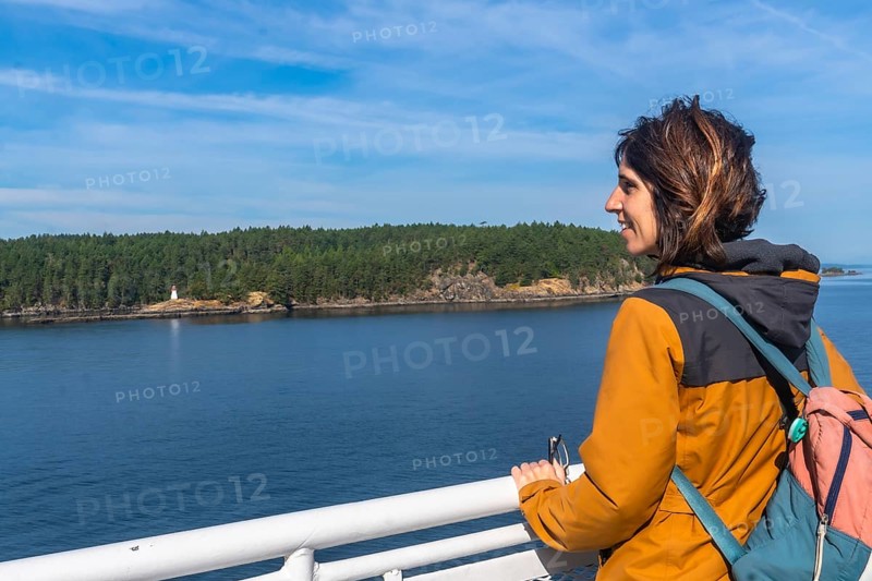 Tourist leaning on the ferry railing, enjoying breathtaking ocean views and a small island with a lighthouse during a scenic journey from victoria to vancouver