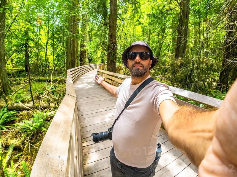 Photographer with hat and sunglasses taking a selfie while walking down a wooden walkway in a lush green forest, enjoying the tranquility of nature and capturing memories