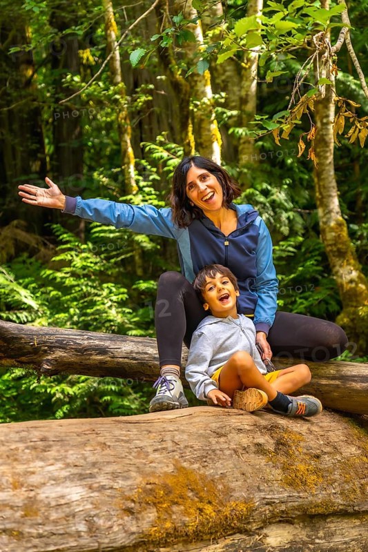Cheerful mother and her son are sitting on a large fallen log in cathedral grove forest on vancouver island, british columbia, canada, enjoying the beauty of nature during summer
