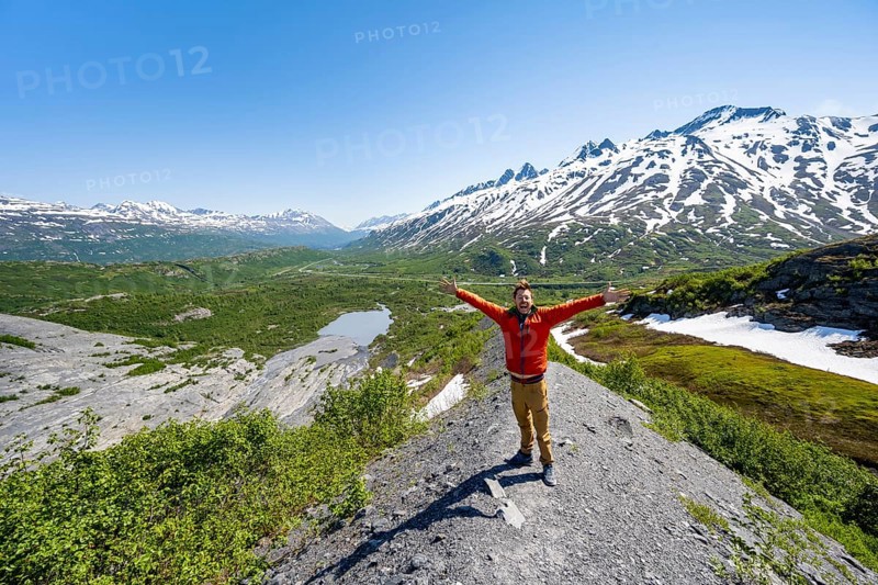 Hiker stretches his arms in the air, on the glacial moraine, view over the wide valley of the Tsina River with mountains, Worthington Glacier State Recreational Site, Chugach Mountains, Alaska, USA