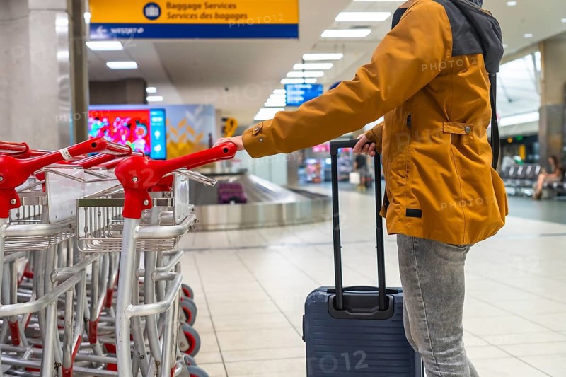 Tourist is pulling his trolley suitcase and taking a luggage cart at the airport terminal, near the baggage claim area, getting ready for his trip