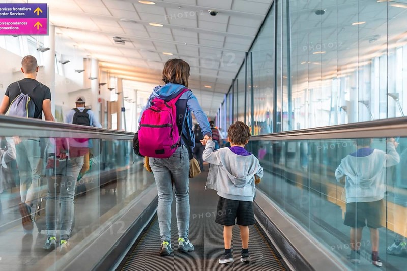 Mother and son with backpacks holding hands walking on a moving walkway in a modern airport terminal, heading towards border services and connecting flights, surrounded by other passengers