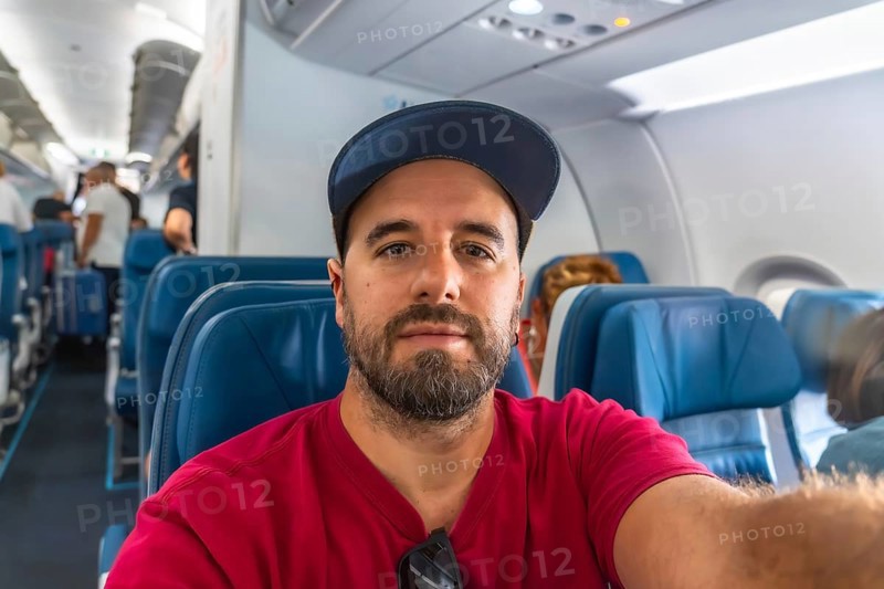 Bearded man capturing a selfie while seated in an airplane during a flight, with fellow passengers boarding in the background, embodying the excitement of travel and adventure
