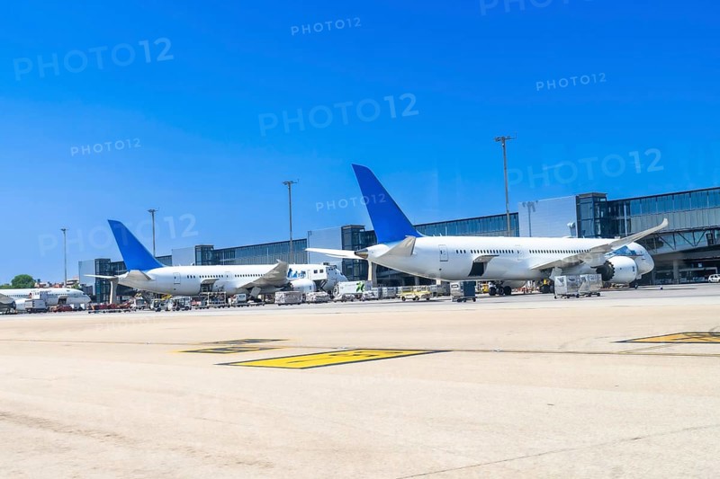 Two large passenger airplanes are parked at the gate of a modern airport on a sunny day, undergoing maintenance and preparation for their next flights