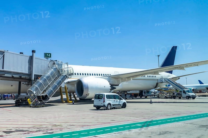 Modern passenger airplane parked at airport gate, connected to jet bridge, with ground crew and support vehicles preparing for passenger boarding on a sunny day