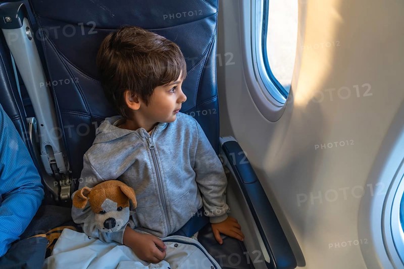 Curious child traveling by plane, clutching a plush toy while gazing out the window, marveling at the expansive sky and fluffy clouds during an exciting flight adventure
