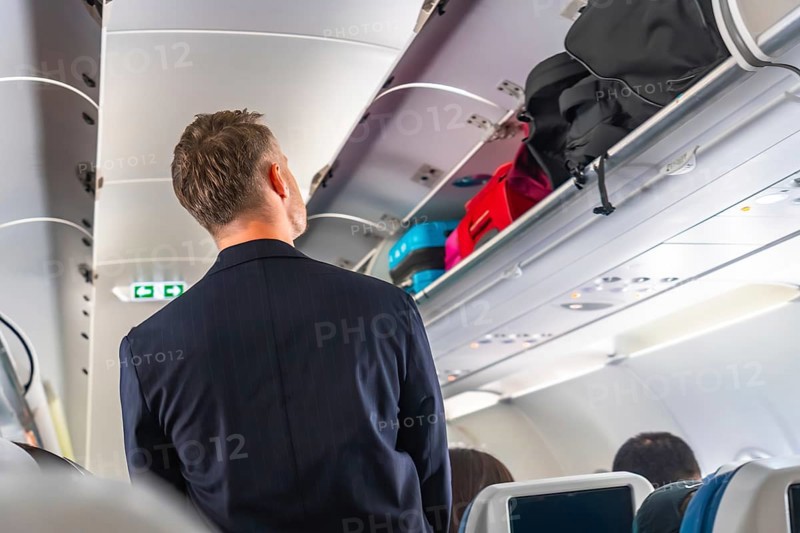 Businessman searching for space to store carry on luggage in the overhead compartment of a crowded passenger airplane during a busy flight, navigating the challenges of air travel