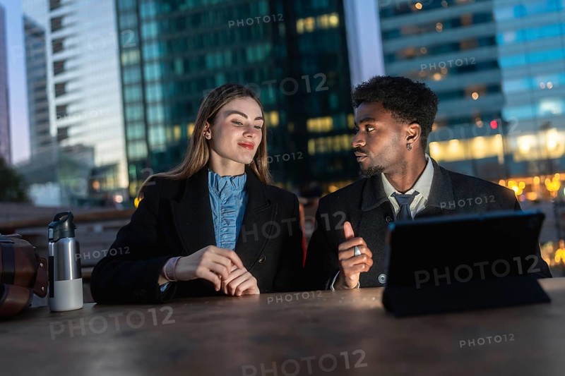 Business colleagues of different ethnicities are sitting at a table with a tablet, engaging in a focused discussion outdoors against the backdrop of modern city buildings at dusk