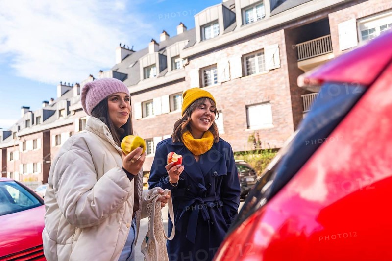 Two young adult women friends enjoying a healthy snack of apples while walking on a city street, embracing a sustainable and active urban lifestyle in a cheerful moment