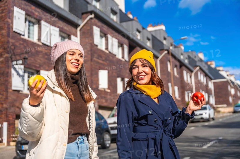 Two smiling young women friends are enjoying a sunny winter day, walking along a city street and holding fresh apples, promoting healthy eating and an active urban lifestyle