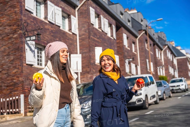 Two cheerful young women enjoying a healthy snack of apples while walking and smiling on a city street during autumn, embracing a vibrant urban lifestyle