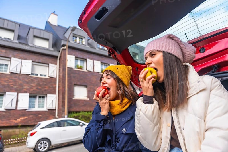 Two young women sit in an open car trunk on a city street, laughing and sharing fresh apples during a casual autumn road trip, enjoying a healthy, carefree moment together