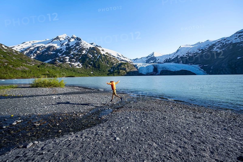 Young man jumping on a pebble beach on a glacial lake, Snowy Mountains and Portage Glacier on Portage Lake glacial lake, Chugach National Forest, Alaska, USA