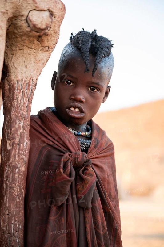 Himba girl leaning at a traditional clay hut, in the morning, traditional Himba village, Kaokoveld, Kunene, Namibia