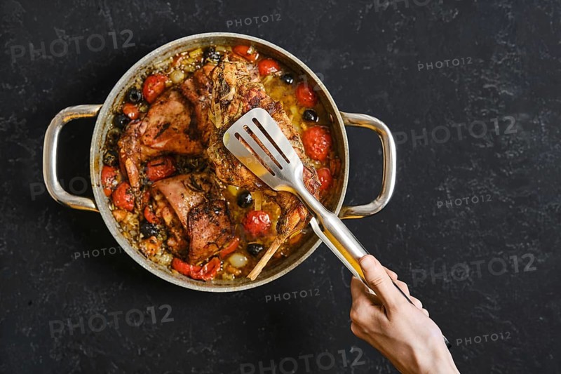 A hand uses a kitchen tongs to lift a tender rabbit leg from a pan filled with roasted tomatoes and olives. This dish is a delightful centerpiece for a cozy meal, rich in flavors and colors