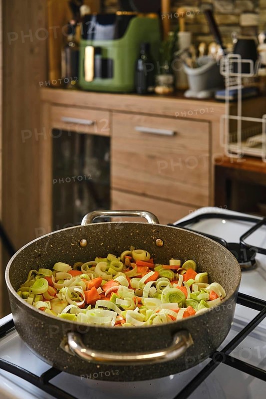 Chopped leeks and carrots sizzle in a large pot on a stovetop. The kitchen features wooden cabinets, modern appliances, and a warm atmosphere, suggesting a cozy cooking experience