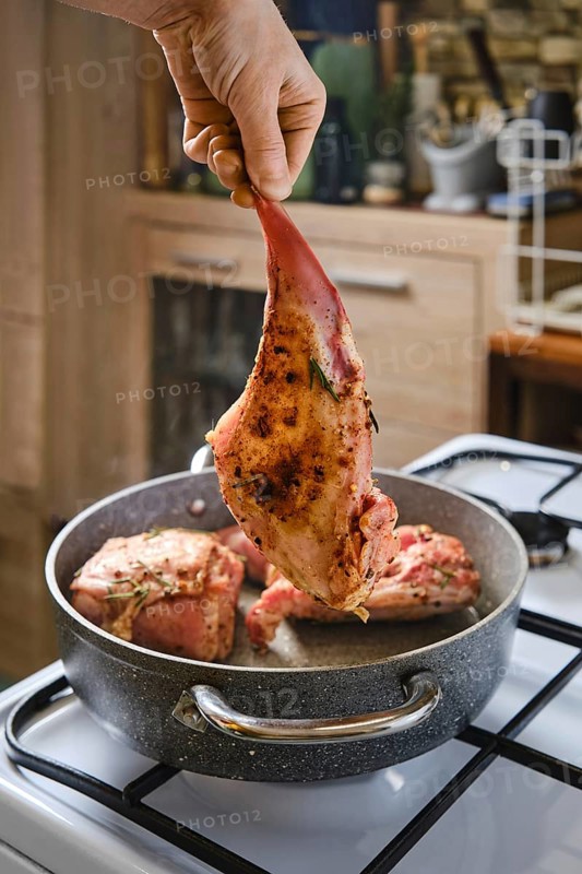 A person holds a marinated rabbit hind leg above a pot with other meat cooking inside. The kitchen has wooden cabinets and a warm atmosphere, indicating a cozy meal preparation time