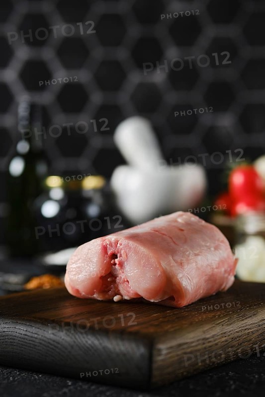 Close-up view of a piece of raw rabbit meat on a wooden cutting board in a contemporary kitchen. Surrounding ingredients include herbs, spices, and kitchen tools, highlighting the preparation for cooking